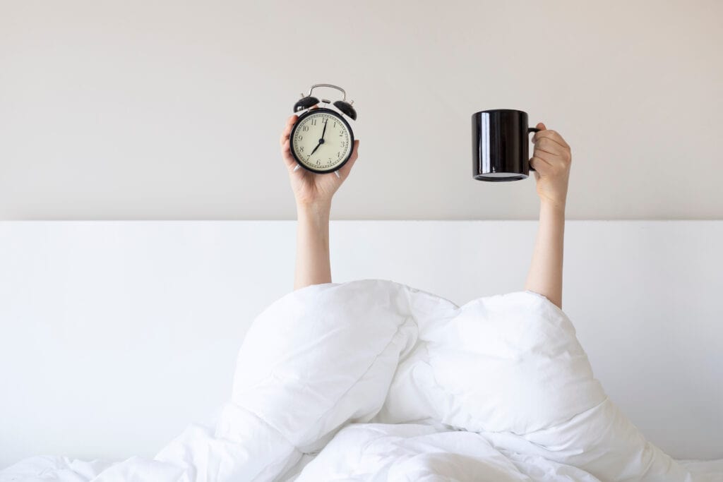 Woman showing arm raised up holding coffee cup and black alarm clock behind duvet in the bed room, Young girl with two hands sticking out from the blanket. wake up with fun in morning concept.