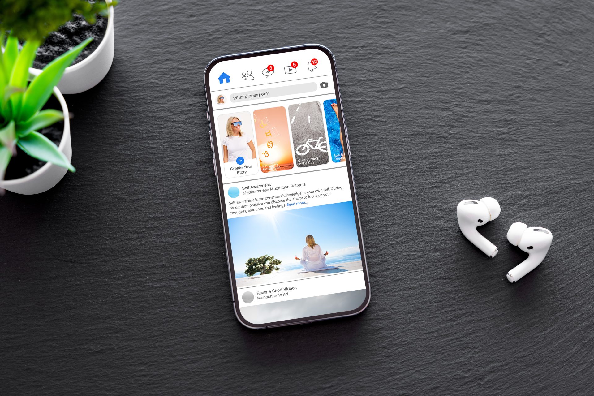Smartphone on a dark slate surface showing a social media feed with notifications, stories, and a meditation post, with a potted plant and wireless earbuds nearby.