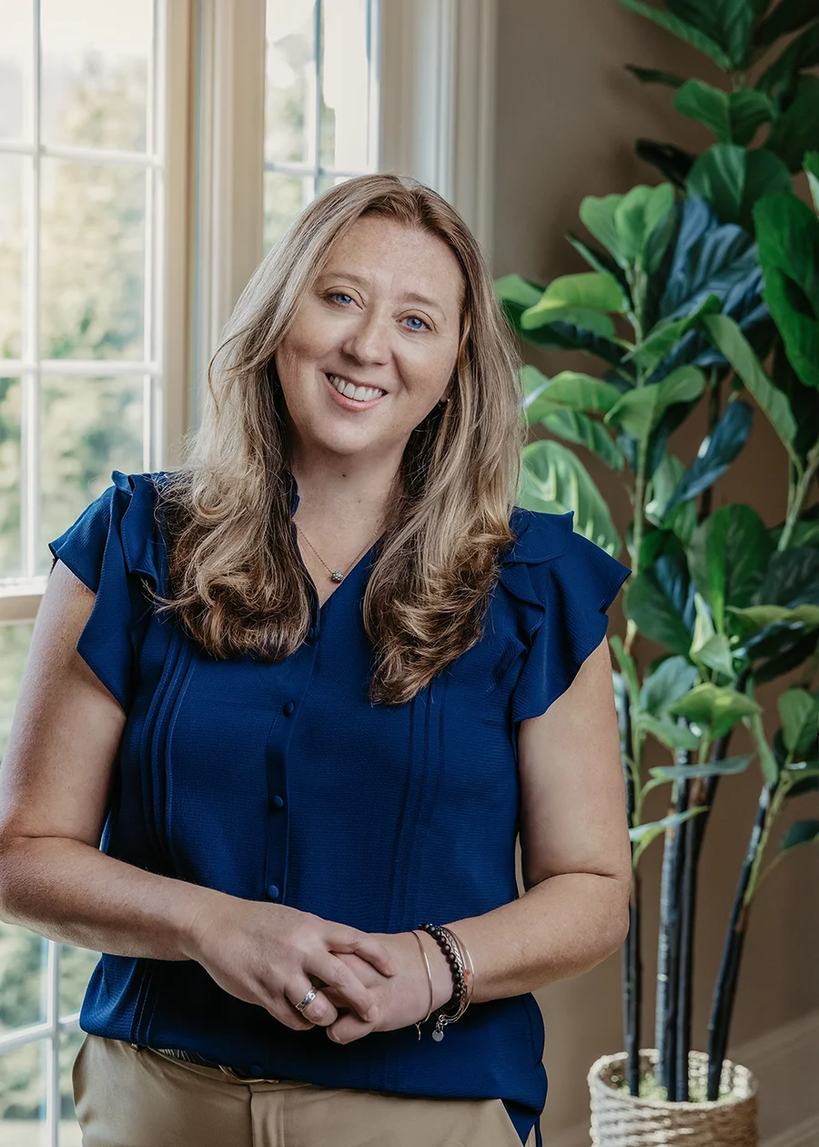 Photo of Heather Johnson wearing a blue top standing next to a window with a plant in the background.
